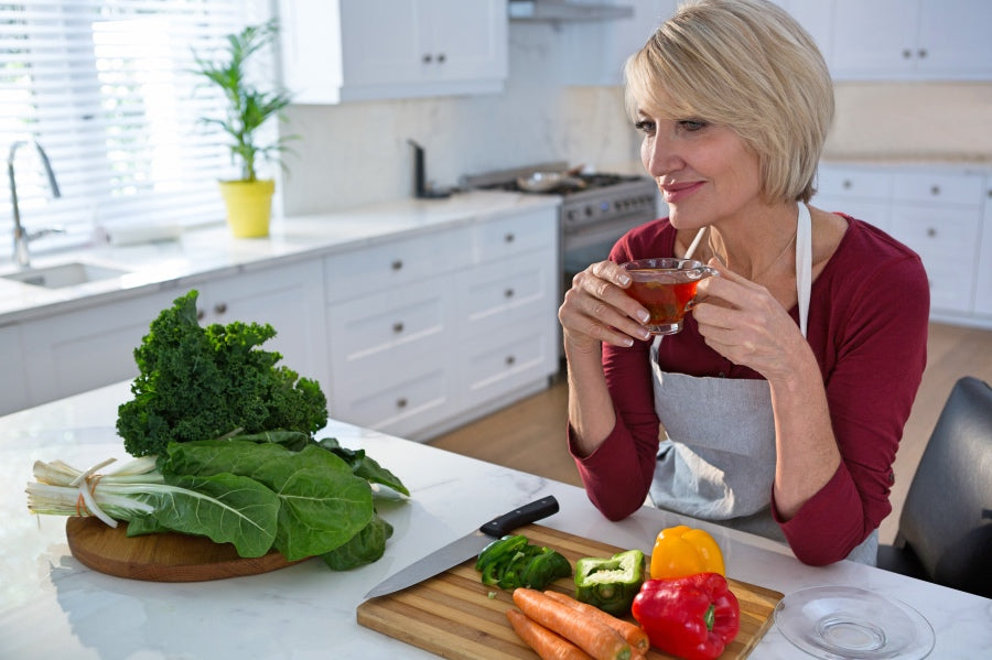 Woman drinking tea at a counter with vegetables and cutting board 