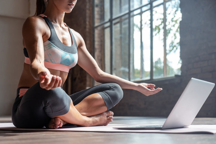 woman sitting with legs crossed on yoga mat in front of laptop learning about meditation benefits