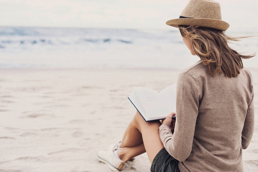A woman sitting on the beach reading with a fedora on